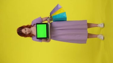 Kızıl saçlı kadın elinde yeşil ekranlı bir laptop tutuyor. Stüdyoda sarı arka planda alışveriş torbaları olan uzun boylu bir kadın. Dikey video. Reklam alanı, çalışma alanı. Satış, Kara Cuma, indirim.