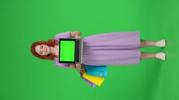 红头发的女人拿着带有绿色屏幕的笔记本电脑 全长的女人在工作室的绿色屏幕上购物袋 垂直录像 广告区 工作区 黑色星期五打折 — 图库照片