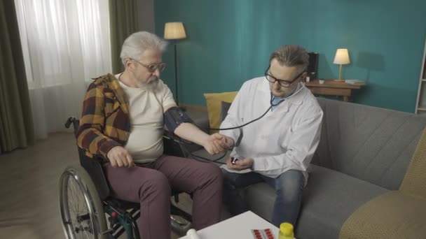 自宅で患者をチェックする医師のフルサイズのビデオ 車椅子の上の高齢者の血圧を測定するためのステートスコープと装置を使用しています メディカルサービス Hdrについて — ストック動画