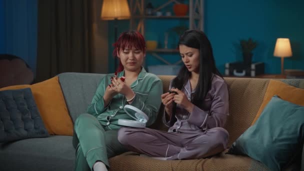 中完整的视频记录了两个穿着睡衣坐在房间沙发上的年轻女性 比较了两种不同的口红 并尝试了其中一种 女孩之夜 — 图库视频影像