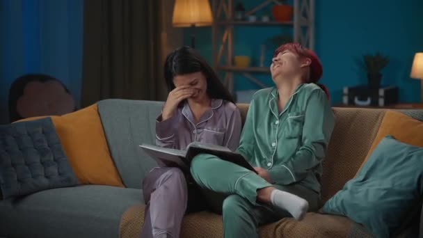 中完整的视频记录了两个穿着睡衣坐在沙发上的年轻女性 翻阅相册 回忆她们的童年 女孩之夜 过夜者 兄弟姐妹 — 图库视频影像