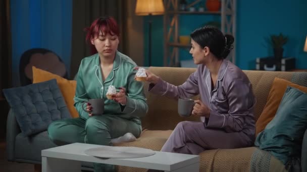 视频中 两个穿着睡衣的年轻女性坐在沙发上 聊着什么 喝着咖啡 吃着纸杯蛋糕 女孩之夜 过夜者 兄弟姐妹 — 图库视频影像