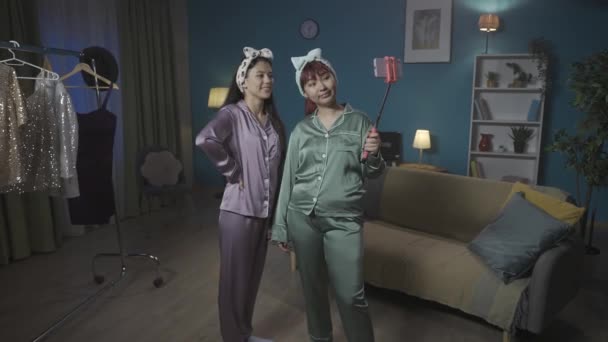 全尺寸的视频记录了两名年轻女性在智能手机上使用自拍棒在房间里拍照的情景 背景上挂着稀奇古怪的服装 留宿者 兄弟姐妹 — 图库视频影像