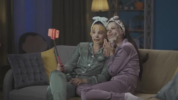 ソファーに座り スキンケアのルーチンを行い セルフィーをとり フェイスマスクを着用したパジャマの2人の若い女性を撮影した中フルビデオ ガールズナイト 睡眠オーバー — ストック動画