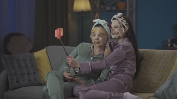 ソファーに座り スキンケアのルーチンを行い セルフィーをとり フェイスマスクを着用したパジャマの2人の若い女性を撮影した中フルビデオ ガールズナイト 睡眠オーバー — ストック動画