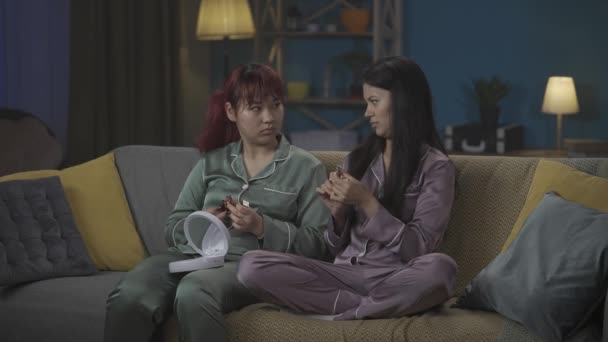 部屋のソファーに座っているパジャマを着た2人の若い女性を撮影した中規模のビデオ 2つの異なる口紅の色合いを比較して そのうちの1つを試してみました ガールズナイト 兄弟姉妹 — ストック動画