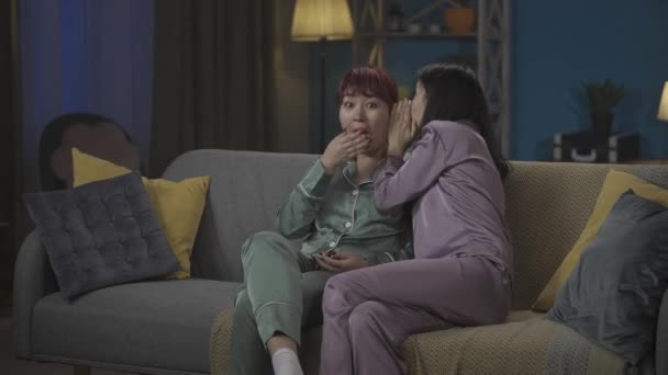 中完整的视频记录了两个穿着睡衣的年轻女性坐在房间沙发上的情景 其中一个和她的朋友 姐姐分享一个秘密的闲话 女孩之夜 过夜者 兄弟姐妹 — 图库视频影像