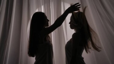 Havluya sarılmış iki kız kardeş, arkadaş, genç kadın silueti. Bir tanesi diğerinin saçlarını yumuşak bir ışıkta perde arkasında sallıyor. Ürün reklamı