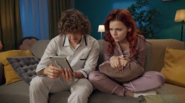 Aile ve ilişkiler reklam konsepti. Birlikte zaman geçiren genç bir çiftin portresi. Pijamalı bir adam ve bir kadın kanepede oturmuş, tablette korku filmi izliyorlar, kız korkuyor..