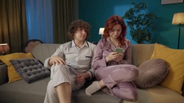 Aile ve ilişkiler reklam konsepti. Birlikte zaman geçiren genç bir çiftin portresi. Pijamalı bir adam ve kadın kanepede oturuyor, kız üzgün, akıllı telefon okuyor, adam ona sarılıyor..