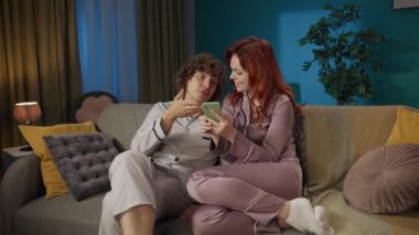 Aile ve ilişkiler reklam konsepti. Birlikte zaman geçiren genç bir çiftin portresi. Pijamalı bir adam ve bir kadın kanepede oturuyor, kız akıllı telefon ekranını erkeklerden saklıyor ve gülüyor..