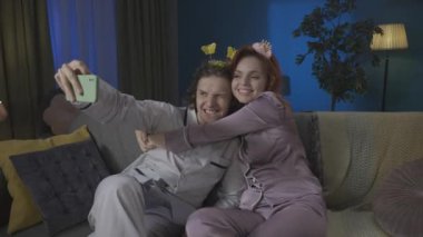 Aile ve ilişkiler reklam konsepti. Birlikte zaman geçiren genç bir çiftin portresi. Pijamalı bir adam ve kadın kanepede sevimli saç bantlarıyla oturuyor ve akıllı telefondan selfie çekiyor. HDR