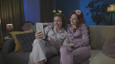 Aile ve ilişkiler reklam konsepti. Birlikte zaman geçiren genç bir çiftin portresi. Pijamalı bir adam ve kadın, tatlı saç bantlarıyla kanepede oturuyorlar ve tabletteki video konuşmalarıyla konuşuyorlar. HDR