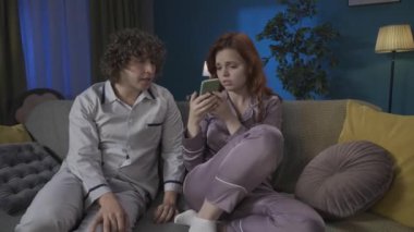 Aile ve ilişkiler reklam konsepti. Birlikte zaman geçiren genç bir çiftin portresi. Pijamalı bir adam ve kadın kanepede oturuyor, kız internette kötü bir şey görüyor, adam onu sakinleştirmeye çalışıyor. HDR