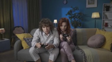 Aile ve ilişkiler reklam konsepti. Birlikte zaman geçiren genç bir çiftin portresi. Pijamalı bir adam ve kadın kanepede oturuyor, video oyunları oynuyor ve şakayla karışık kavga ediyor, kız kazandı. HDR