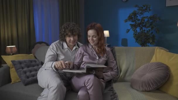 家族と人間関係の広告コンセプト 一緒に過ごした若いカップルの肖像画 ソファに座っているパジャマの男女 古い写真アルバムを見て 笑って 写真を隠す — ストック動画
