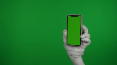 Detaylı yeşil ekran izole edilmiş krom anahtar videosu. Bir mumyanın elinde reklam alanı olan akıllı bir telefon var. Çalışma alanı tanıtım videonuz için maket yapın ve ekranı tıklatın..