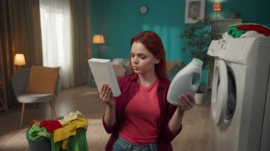 Kızıl saçlı kadın çamaşır makinesi ve çamaşır sepetinin yanında duruyor, iki deterjan arasında seçim yapıyor, toz haline gelmiş olan yerine sıvı deterjanı seçiyor. Ev aletleri, ev işleri, reklam..
