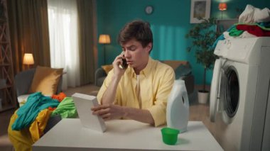 Koyu renk saçlı bir adam çamaşır makinesinin yanında oturuyor, telefonda biriyle konuşuyor. Böylece toz ve sıvı deterjan arasında seçim yapmasına yardım edebileceklerdi. Ev aletleri, ev işleri, reklam.