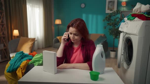 红头发女人坐在洗衣机旁边 与人通电话 这样他们就能在粉末和液体洗涤剂之间做出选择 家用电器 家务活 — 图库视频影像