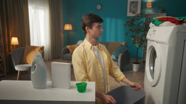Çamaşır makinesinin yanında oturan, elinde boş bir sepet tutan ve programın çalışmasını izleyen siyah saçlı adam. Ev aletleri, ev işleri, reklam..