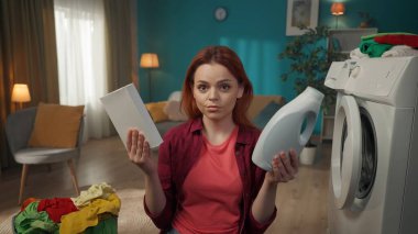 Çamaşır makinesi ve çamaşır sepetinin yanında duran kızıl saçlı kadın toz ve sıvı deterjan arasında seçim yapıyor, neyi seçeceğini şaşırmış. Ev aletleri, ev işleri, reklam..