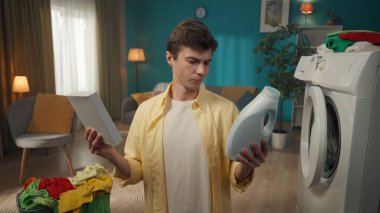 Çamaşır makinesi ve çamaşır sepetinin yanında oturan koyu renk saçlı adam toz ve sıvı deterjan arasında seçim yapıyor, neyi seçeceğini şaşırıyor. Ev aletleri, ev işleri, reklam..