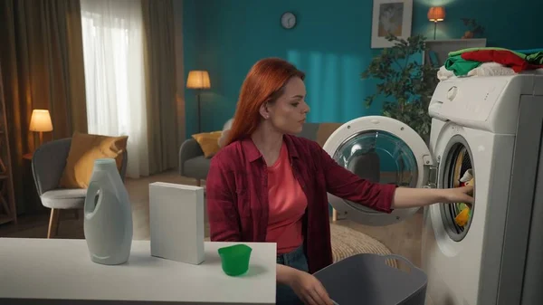 红头发女人站在桌子旁边 拿着洗涤剂 在洗衣机里或从洗衣机里装卸衣服 家用电器 家务活 促销和广告 — 图库照片