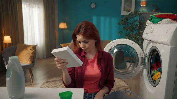 红头发的女人坐在一台洗衣机旁边 这台洗衣机刚刚用完后 把说明书读给粉末状洗涤剂听 家用电器 家务活 — 图库照片