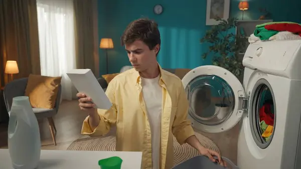 一个黑头发的男人坐在一台洗衣机旁边 这台洗衣机刚刚用完 他把说明书读给一个粉状洗涤剂听 家用电器 家务活 — 图库照片