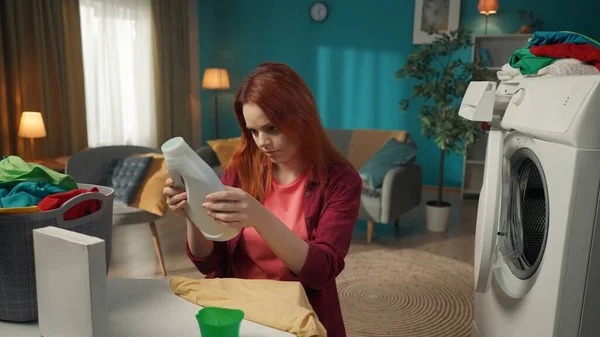 红头发的女人坐在洗衣机旁边 手里拿着一篮子衣服 一边读说明书 一边看液体洗涤剂 家用电器 家务活 — 图库照片