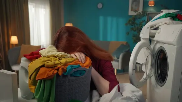 Redhead woman sitting next to a washing machine, laying her head on a laundry basket, frustrated and exhausted about the household chores she has to do. Household appliances, chores, advertisement.