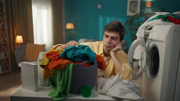 Dark-haired man sitting next to a washing machine, looking at the laundry basket, exhausted about the household chores she has to do. Household appliances, chores, advertisement.