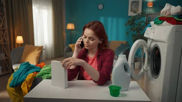 红头发女人坐在洗衣机旁边 与人通电话 这样他们就能在粉末和液体洗涤剂之间做出选择 家用电器 家务活 — 图库照片