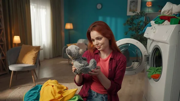 红头发的女人站在房间里 卸下洗衣机 兴奋而高兴地发现一只泰迪熊在那里 充满爱心地看着它 家用电器 家庭价值观 家务活 — 图库照片