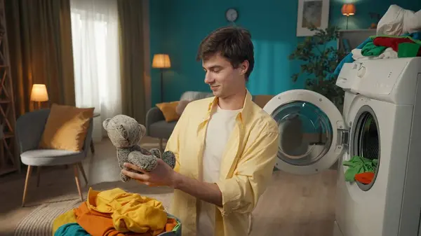 黑头发的男人站在房间里 卸下洗衣机 兴奋地 高兴地看到一只泰迪熊 充满爱心地看着它 家用电器 家庭价值观 家务活 — 图库照片