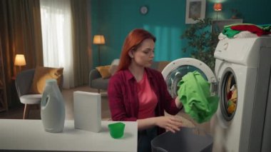 Çamaşır makinesinin yanında duran kızıl saçlı kadın, yıkanmayan giysileri boşaltıyor ve deterjanla kutuya bakıyor, sonuçtan bıkmış. Ev aletleri, ev işleri, reklam..