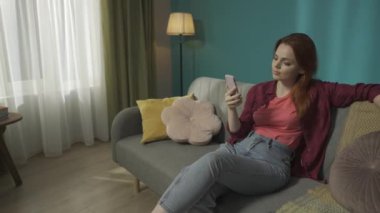 Kanepede oturan, sohbet eden, telefonda bir şey izleyen kızıl saçlı bir kadının ön planda çalışan çamaşır makinesini gösteren videoyu büyüt. Ev aletleri, ev işleri, reklam.