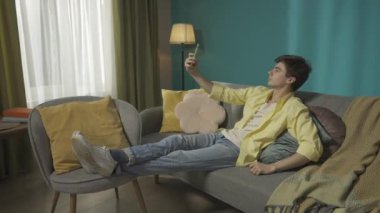 Koyu renk saçlı bir adamın kanepeye oturup selfie çekerken, telefonda sohbet ederken videoyu büyüt. Çamaşır makinesi ön planda çalışıyor. Ev aletleri, ev işleri, reklam.