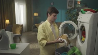Koyu renk saçlı bir adam çamaşır makinesinin yanında duruyor, yıkanmayan giysileri boşaltıyor, kameraya lekeli bir tişört gösteriyor, başını sallıyor. Ev aletleri, ev işleri, reklam. HDR