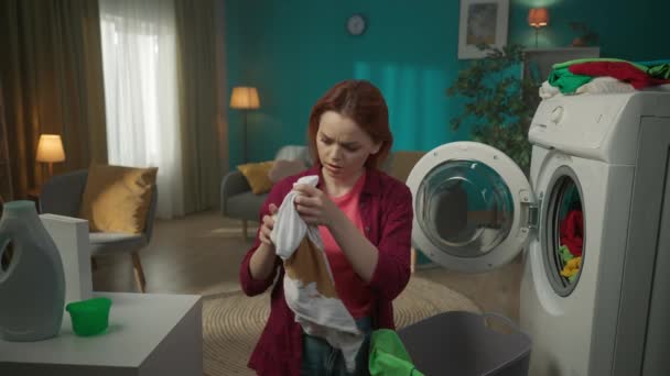 红头发的女人站在洗衣机旁边 卸下洗不掉的衣服 在相机前展示一件污迹斑斑的T恤 摇着头 家用电器 家务活 — 图库视频影像