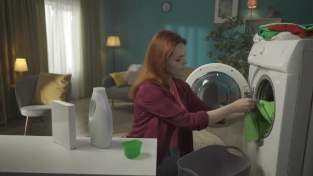 红头发的女人站在洗衣机旁边 卸下干净的衣服 闻到它们的味道 用洗涤剂看着瓶子 兴奋地看到结果 家用电器 家务活 — 图库视频影像
