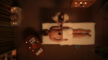 Vücut bakımı reklam konsepti. Prosedür uygulanan kadın portresi. Spa salonunun en iyi manzarası, masaj masasındaki kadın, kadın terapist sırtına siyah masaj taşları koyuyor. Yavaş çekim.