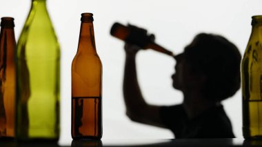 Beyaz arka planda çok renkli boş şişelerin yakın çekimi. Arka planda bir adamın silueti var. Kameraya yan yatmış, şişeyi üzerine kaldırmış, ama boş..