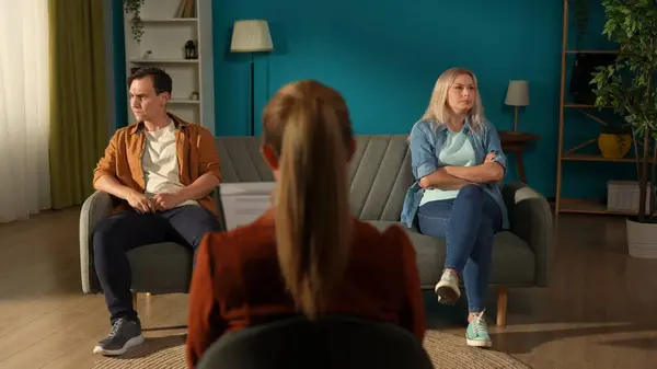 Married couple sitting on different sides of the couch in front of the female therapist. A man and a woman in a quarrel avoid eye contact. Crisis in relationships