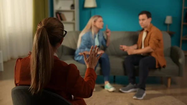 Back view of a female therapist, against a blurred background of a couple arguing. The therapist is trying to calm a man and woman blaming each other for problems. A married couple sitting on a couch