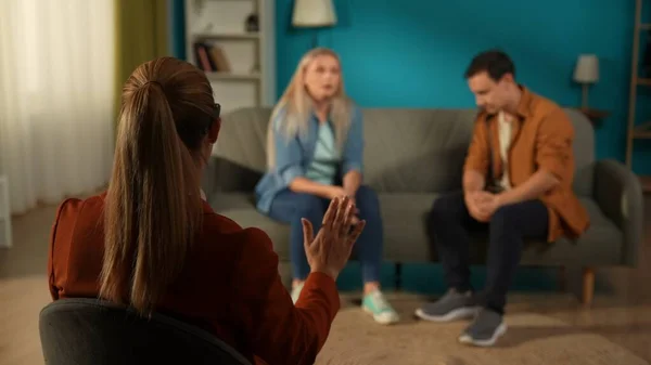 Back view of a female therapist, against a blurred background of a couple arguing. The therapist is trying to calm a man and woman blaming each other for problems. A married couple sitting on a couch