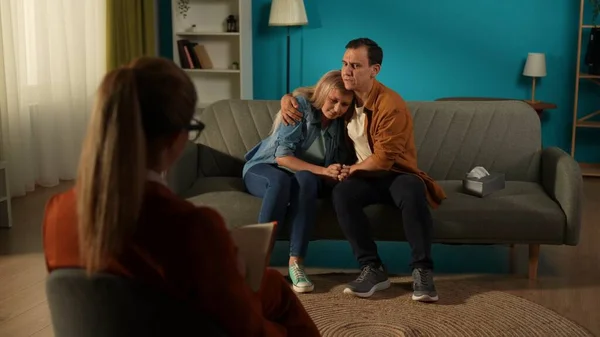 A married couple in a couples therapy session, sitting on a couch in front of a female counselor. A loving attentive man, hugs and comforts the crying spouse. The young woman suffers from depression