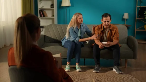 A married couple in a couples therapy session, sitting on a couch in front of a female counselor. A loving attentive woman hugs and comforts a crying spouse. A young man suffers from depression