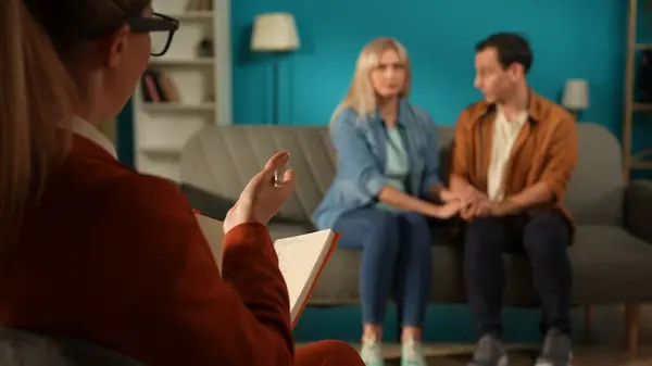Family counseling. Married couple holding hands sitting on a couch in front of a female psychologist. Back view of a female psychologist with a clipboard in her hand